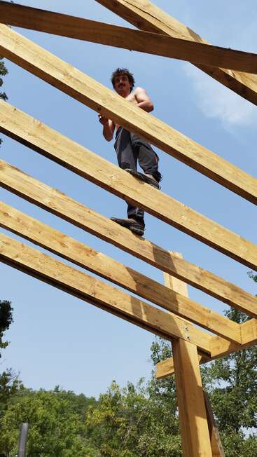 Un hombre sin camiseta sobre las vigas de madera de un tejado en construcción, en equilibrio sobre el suelo, con un cielo azul brillante de fondo.
