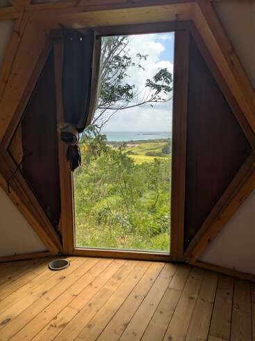 Ein sechseckiges Fenster mit Holzrahmen bietet einen malerischen Blick auf grüne Felder, Bäume und das ferne Meer unter einem teilweise bewölkten Himmel, während Sonnenlicht ins Innere strömt.