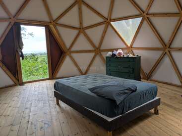 Dieses Bild zeigt ein minimalistisch gestaltetes, kuppelförmiges Schlafzimmer mit geometrischen Holzelementen, einem schlichten Bett mit grauer Bettwäsche, einer grünen Kommode, einem Holzboden und einer offenen Tür, durch die man einen malerischen Ausblick hat.