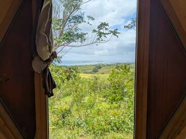 Durch ein Fenster mit Holzrahmen erstreckt sich eine üppig grüne Landschaft bis hin zu fernen Hügeln und einem blauen Meer unter einem teilweise bewölkten Himmel, was ein Gefühl von Ruhe und natürlicher Schönheit vermittelt.