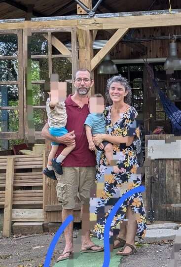 Eine vierköpfige Familie steht vor einem rustikalen Holzhaus. Der Vater und die Mutter tragen jeweils ein Kind auf dem Arm. Einige Teile des Bildes sind verpixelt.