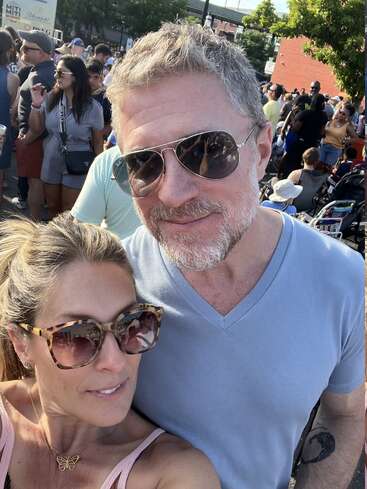 The image shows a selfie of a man and woman at an outdoor event, with a crowd of people in the background, likely a festival or concert.
