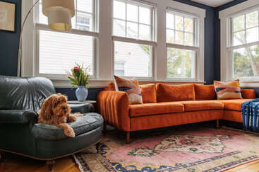 The image depicts a living room with a couch, coffee table, and bookshelf, featuring a large window and a fireplace, with a neutral color scheme and a minimalist aesthetic.
