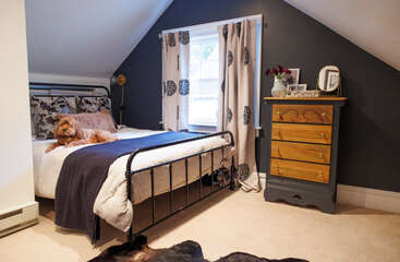 The image depicts a cozy room with a bed, dresser, and desk, featuring a dog in the background, set against a neutral-colored wall and floor. The overall atmosphere is one of warmth and comfort.