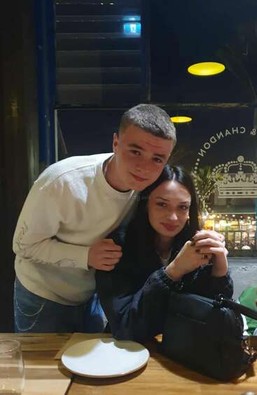 Un joven y una joven posan juntos en la mesa de un restaurante. Ella está sentada con las manos entrelazadas, mientras él se inclina hacia ella; ambos sonríen amablemente a la cámara.