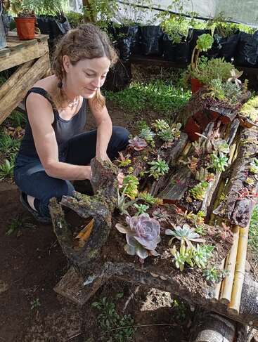 A woman in a black tank top tends to a rustic garden bed filled with various succulents, surrounded by greenery inside a shaded outdoor greenhouse.
