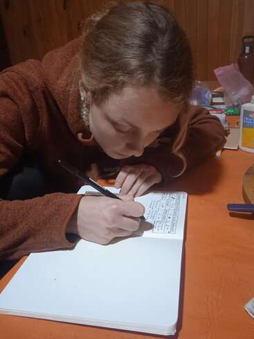 A woman with light brown hair, wearing a brown sweater, is intently writing in a notebook at a wooden table with stationery items scattered around.