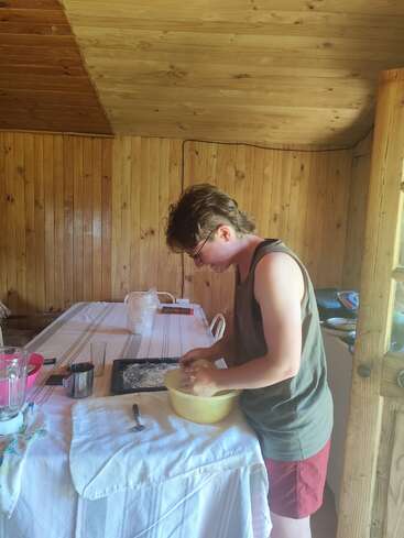 A person mixes ingredients in a yellow bowl on a table covered with a white cloth inside a wooden cabin kitchen, preparing for baking or cooking.