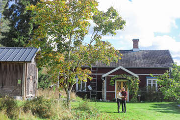 Una pareja se abraza sobre un césped verde frente a una encantadora casa de madera roja, rodeada de árboles y luz solar, creando un apacible ambiente campestre.