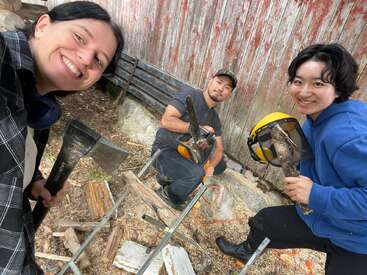 Tres personas sonrientes están al aire libre cortando leña; una sostiene un hacha, otra maneja una motosierra y la tercera sostiene la leña y el equipo de seguridad. Entorno rústico, trabajo en equipo.