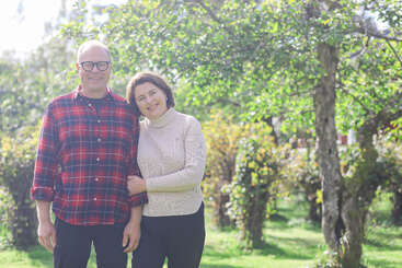 Una pareja sonriente permanece unida al aire libre en un jardín soleado y verde. La mujer se apoya en el hombro del hombre. Los árboles y la exuberante vegetación les rodean apaciblemente.
