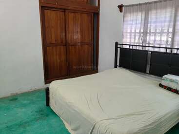 Um quarto simples com uma cama, lençóis brancos, toalhas dobradas, um guarda-roupa de madeira, carpete verde e uma grande janela com cortinas transparentes que deixam entrar a luz do dia.