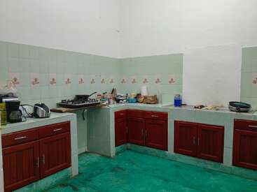 Esta é uma cozinha modesta com paredes de azulejos verdes, armários vermelhos, piso verde, um fogão, vários eletrodomésticos, utensílios, ingredientes e decoração mínima, criando um espaço funcional.