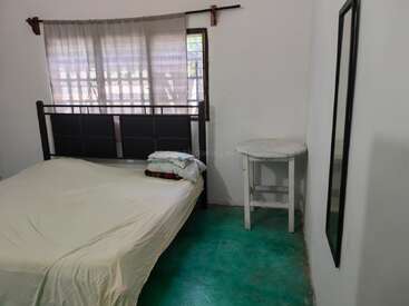 Este é um quarto simples com uma cama, travesseiros empilhados, piso verde, janela com cortinas, mesa redonda branca e um espelho de parede alto.