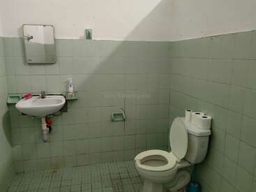 Esse é um banheiro básico, pouco mobiliado, com azulejos verdes. Ele contém uma pia, espelho, vaso sanitário aberto, rolos de papel higiênico e um frasco de sabonete.