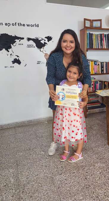 Uma mulher e uma jovem sorrindo, segurando um diploma. A garota usa um vestido de coração. Estantes de livros e um mapa-múndi decoram o plano de fundo.