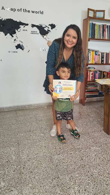 Uma mulher e um menino posam, sorrindo, enquanto seguram um diploma. Atrás deles, há uma estante cheia de livros e um mapa-múndi na parede.