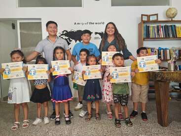 Um grupo de crianças sorridentes exibe orgulhosamente seus diplomas ao lado de dois adultos em uma sala de aula, com estantes, um globo e um mapa-múndi de fundo.