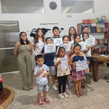 Um grupo de crianças e adultos posa em um ambiente interno, segurando orgulhosamente certificados. Eles estão em frente a uma parede com um mapa-múndi, estantes de livros e sorrindo para a câmera.
