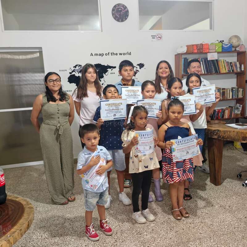 Um grupo de crianças e adultos posa em um ambiente interno, segurando orgulhosamente certificados. Eles estão em frente a uma parede com um mapa-múndi, estantes de livros e sorrindo para a câmera.