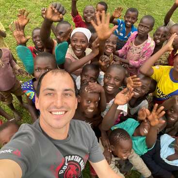 La imagen muestra a un hombre tomándose un selfie con un grupo de niños en un campo cubierto de hierba, todos sonriendo y saludando a la cámara.