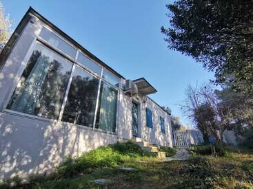 La imagen muestra una casa blanca con grandes ventanales, un pequeño patio y un jardín sereno, bajo un cielo azul despejado.