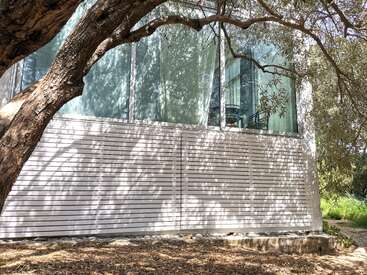 La imagen muestra un moderno edificio blanco con grandes ventanales, un árbol en primer plano y un sereno entorno exterior.