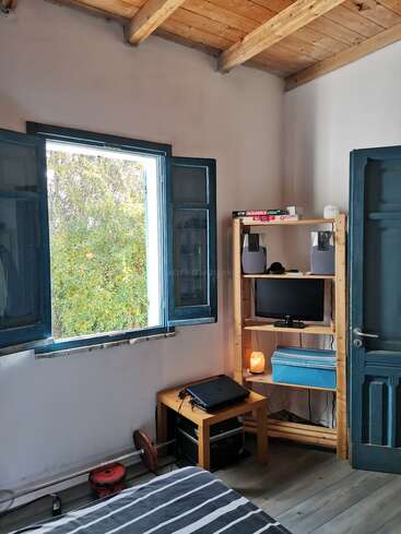 Una acogedora habitación con ventanas y puerta azules, techo de madera, cama a rayas, mesita, estantería con juegos, altavoces, monitor, lámpara y vegetación visible en el exterior.