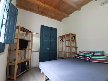 Dieses gemütliche Zimmer verfügt über eine Holzdecke, blaue Flügeltüren, offene Regale, einen Fernseher, gestreifte Kissen, ein Bett, Fensterläden und eine minimalistische, funktionale Einrichtung.