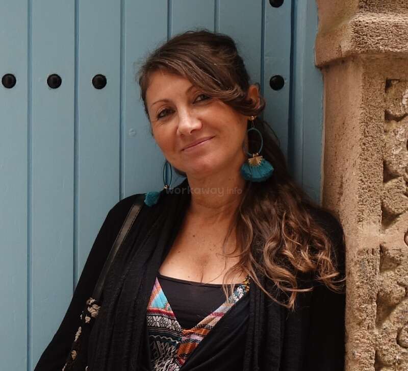 La imagen muestra a una mujer con el pelo largo y castaño, vestida con una rebeca negra y un top de colores, de pie frente a una puerta azul y una pared bronceada, con una sutil marca de agua visible.