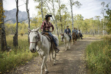 A group of people on horseback ride through a scenic forest trail bordered by tall trees, with mountains in the background. They wear plaid shirts and hats.