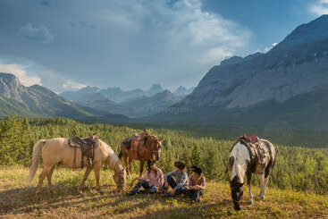 Three people in cowboy hats sit on grass, relaxing with three saddled horses. Majestic mountains, green forests, and a dramatic sky form the breathtaking background.