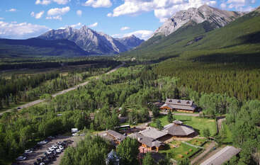 A scenic aerial view shows rustic lodges nestled among dense green forest, with rugged mountains towering in the background under a bright blue sky with clouds.