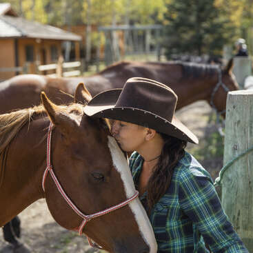 A woman in a cowboy hat kisses a brown horse on its forehead, showing affection. Another horse stands in the background. The scene is outdoors, peaceful.