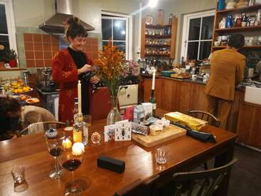 La imagen representa una escena de cocina cálida y acogedora, con una mesa de madera con copas de vino, velas y un jarrón de flores, rodeada de gente disfrutando de una comida.