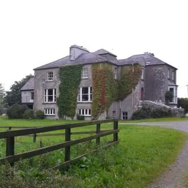 La imagen muestra una gran casa de piedra gris con un frondoso césped verde y una valla de madera, con paredes cubiertas de hiedra y múltiples ventanas.