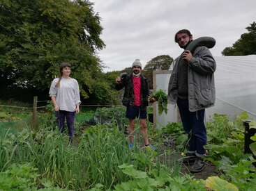 Tres personas de pie en un jardín, rodeadas de exuberante vegetación y plantas, con un cielo nublado por encima, transmitiendo una sensación de comunidad y actividad al aire libre.