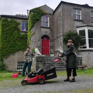 La imagen muestra a dos personas delante de una gran casa de piedra desgastada, una de ellas manejando un cortacésped rojo y negro.