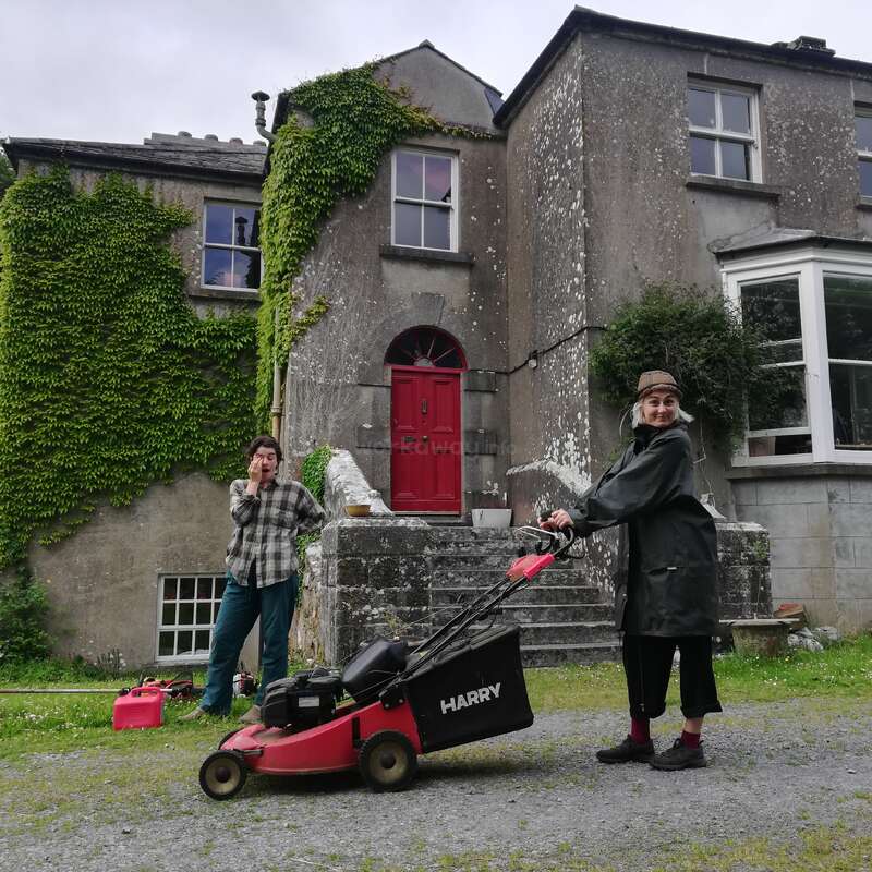 La imagen muestra a dos personas delante de una gran casa de piedra desgastada, una de ellas manejando un cortacésped rojo y negro.