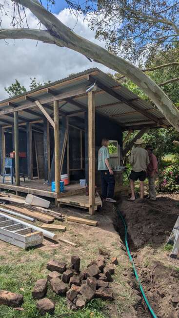 Drei Leute stehen neben einem halbfertigen Holzschuppen. Überall liegen Werkzeuge, Ziegelsteine und Baumaterialien rum, und in der Nähe gibt's einen Graben und einen Garten unter blauem Himmel.