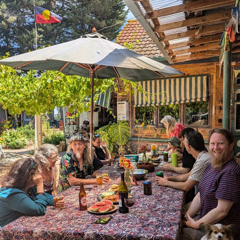 Eine Gruppe Leute genießt ein sonniges Essen im Freien unter einem großen Sonnenschirm, umgeben von viel Grün. Essen, Getränke, Gelächter und ein Hund sorgen für eine fröhliche Stimmung.