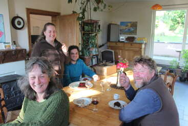 Four people enjoy a cheerful gathering around a wooden dining table with cake and wine. The sunlit room feels cozy, filled with plants and laughter.