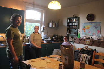 Three people are smiling and talking in a cozy kitchen. Dough and ingredients are on the table, suggesting baking. The room is warmly lit and inviting.