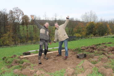 Two people stand joyfully on a muddy patch, surrounded by grass, dirt, and tools, in a scenic field with trees in the background during autumn.