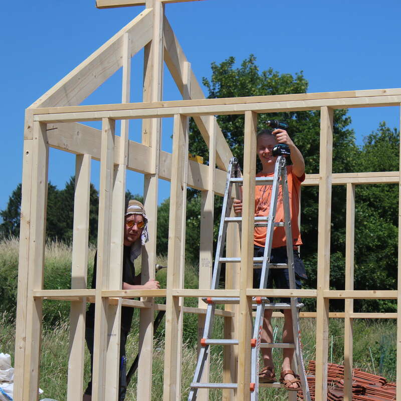 Two people are constructing a wooden frame for a house on a sunny day. One person stands on a ladder, using a drill, while the other observes.