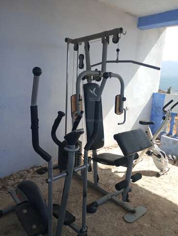 Dieses Bild zeigt Fitnessgeräte, darunter eine Kraftstation, eine elliptische Maschine und einen Heimtrainer. Der Fitnessraum scheint sich im Freien zu befinden, wo das Sonnenlicht hineinfällt.