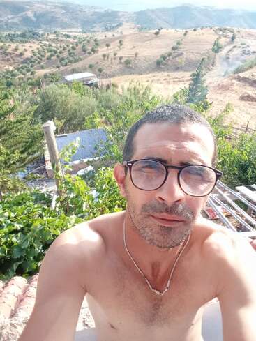 Ein Mann mit Brille und einer Halskette macht ein Selfie im Freien. Er ist hemdsärmelig, mit grünem Laub und sanften Hügeln im sonnigen Hintergrund.