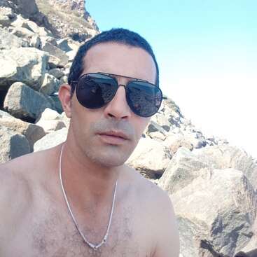 Un hombre con gafas de sol y collar posa para un selfie al aire libre. Está sin camiseta, sentado sobre unas rocas, con un cielo azul despejado a sus espaldas.