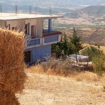 Uma casa azul com um telhado inacabado fica em uma encosta seca. Fardos de feno, árvores verdes e montanhas distantes completam essa pacífica cena de paisagem rural.