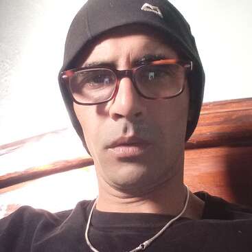 Das Bild zeigt eine Person, die eine schwarze Mütze, eine Brille und ein schwarzes Hemd trägt, mit einer weißen Kordel um den Hals, vor dem Hintergrund einer weißen Wand und einer Holzoberfläche.